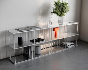 May include: A modern, three-tiered white and silver metal shelf unit. The shelves hold a variety of items, including books, a smart speaker, a tablet, and an orange table lamp. A vase with greenery and the word "project" are visible.