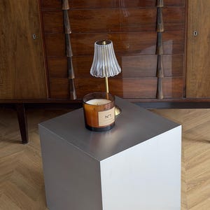 Op de afbeelding: Een moderne, zilverkleurige kubusvormige bijzettafel. Een brandende kaars in een glazen pot met het label "N°1" staat erop, naast een kleine lamp met een gegolfde glazen kap. Op de achtergrond een donkere houten kast.
