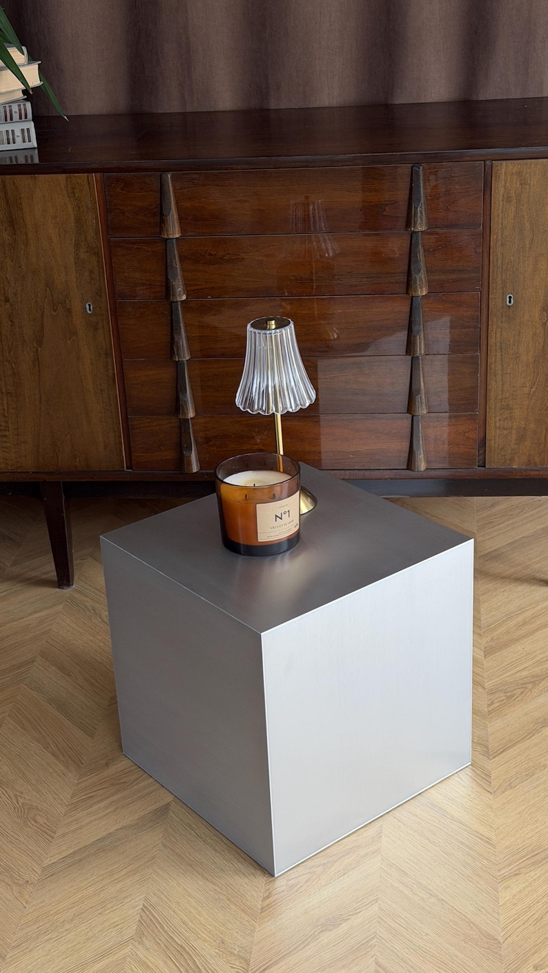 May include: A modern, silver-colored cube-shaped side table. A lit candle in a glass jar with a label that reads "N&deg;1" sits on top, next to a small lamp with a fluted glass shade. A dark wood cabinet is in the background.