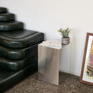 Könnte beinhalten: Ein moderner Beistelltisch mit gebürsteter Metalloberfläche und einer transparenten Acrylplatte. Eine kleine Topfpflanze und ein Buch liegen auf der Tischoberfläche. Der Tisch steht neben einem dunkelgrünen Ledersofa und einem gerahmten Kunstwerk.