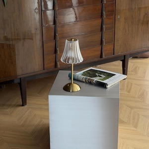 May include: A small, decorative lamp with a clear, ribbed glass shade and a gold-colored base sits on a gray cube-shaped table. A magazine is placed on the table. The background features a wooden cabinet and a parquet floor.
