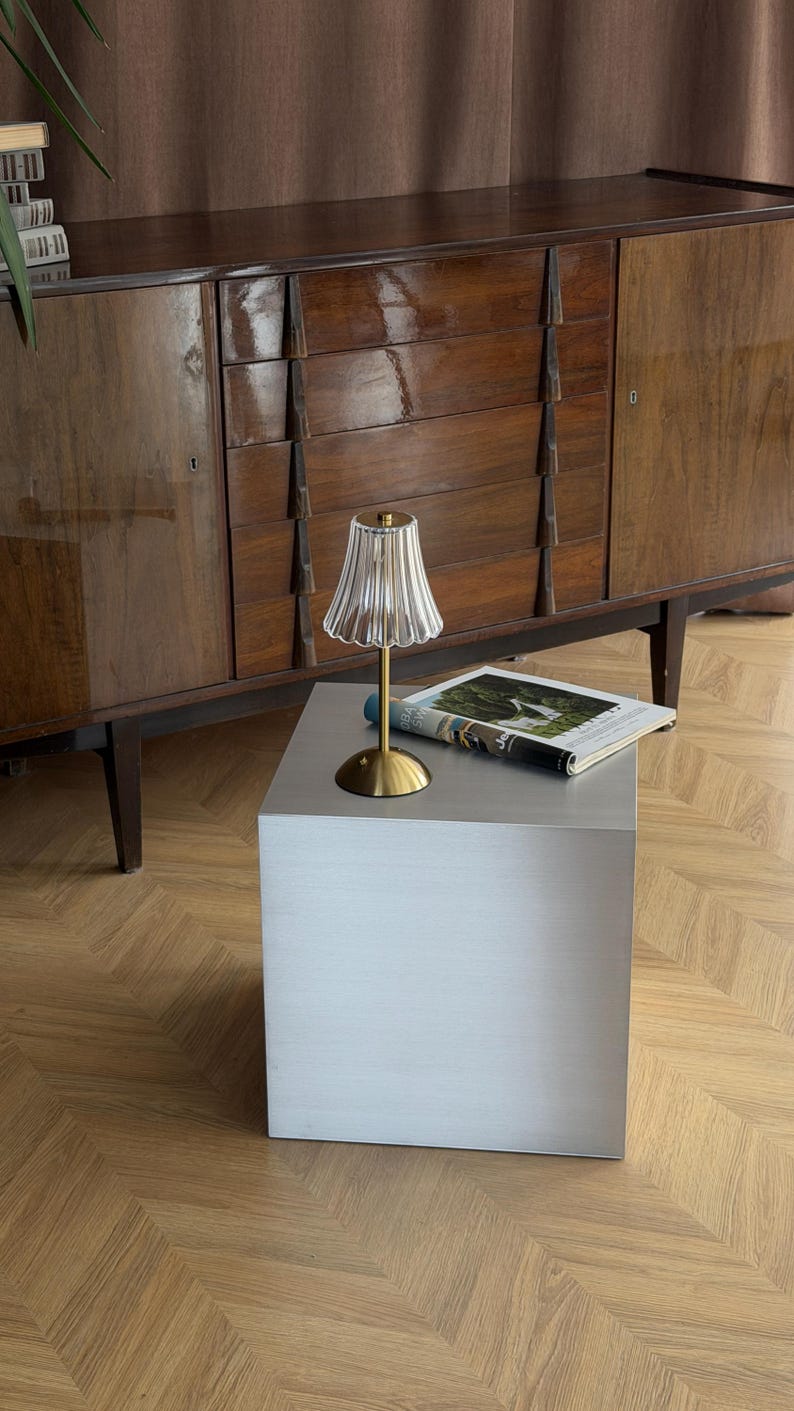 May include: A small, decorative lamp with a clear, ribbed glass shade and a gold-colored base sits on a gray cube-shaped table. A magazine is placed on the table. The background features a wooden cabinet and a parquet floor.