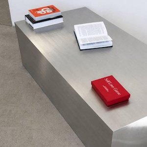 May include: A brushed metal rectangular coffee table with books and a red box. Two books are stacked, one with an orange cover and the other with a black cover. An open book and a red box with the text "Self-Love Game" are also on the table.