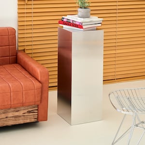 May include: A modern, silver metal pedestal table with a reddish-brown accent panel. The table supports a small potted plant and a stack of books. A leather armchair and a wire chair are also visible.