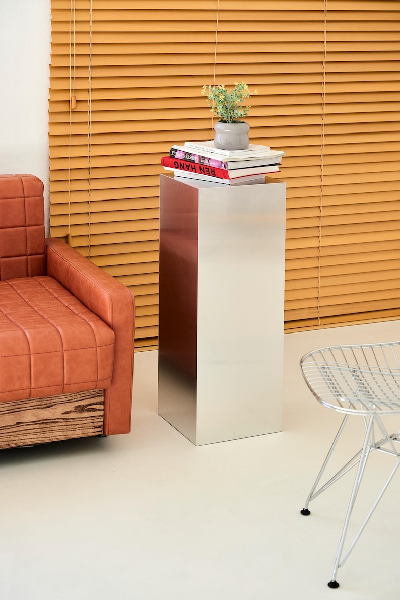 May include: A modern, silver metal pedestal table with a reddish-brown accent panel. The table supports a small potted plant and a stack of books. A leather armchair and a wire chair are also visible.