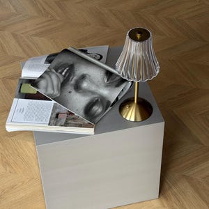 May include: A small, light gray cube table with a brass-colored lamp and a clear, fluted lampshade. Magazines with black and white photography are stacked on the table. The table sits on a wooden floor.
