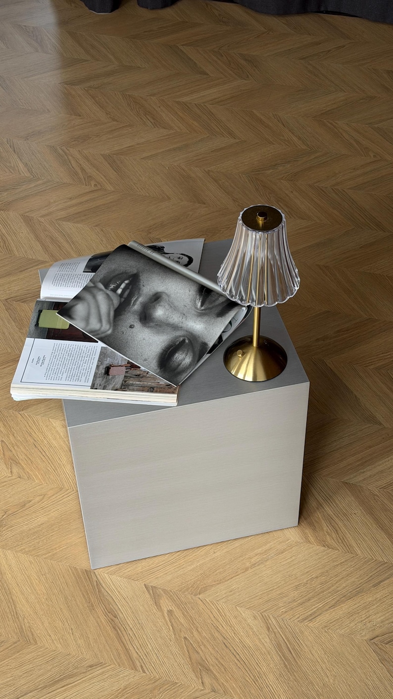 May include: A small, light gray cube table with a brass-colored lamp and a clear, fluted lampshade. Magazines with black and white photography are stacked on the table. The table sits on a wooden floor.