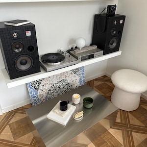 May include: A modern living room setup featuring a silver metal coffee table, a turntable, and black speakers. A black mug, a green bowl, and a candle sit on the table. The room has a wooden floor and white shelves.