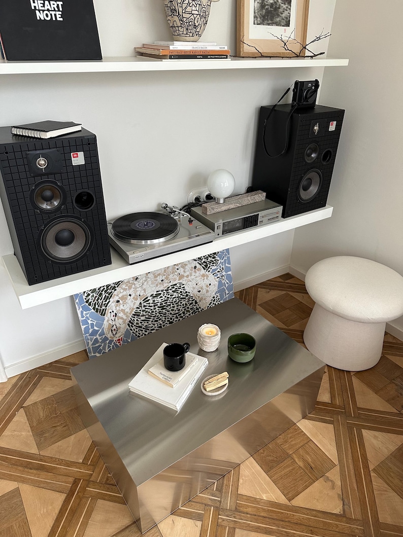 May include: A modern living room setup featuring a silver metal coffee table, a turntable, and black speakers. A black mug, a green bowl, and a candle sit on the table. The room has a wooden floor and white shelves.