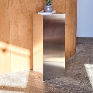 May include: A modern, stainless steel side table with a square top and base. A small, gray ceramic pot with a plant sits on top of the table, resting on a white notepad. The table is in a room with a wooden wall.