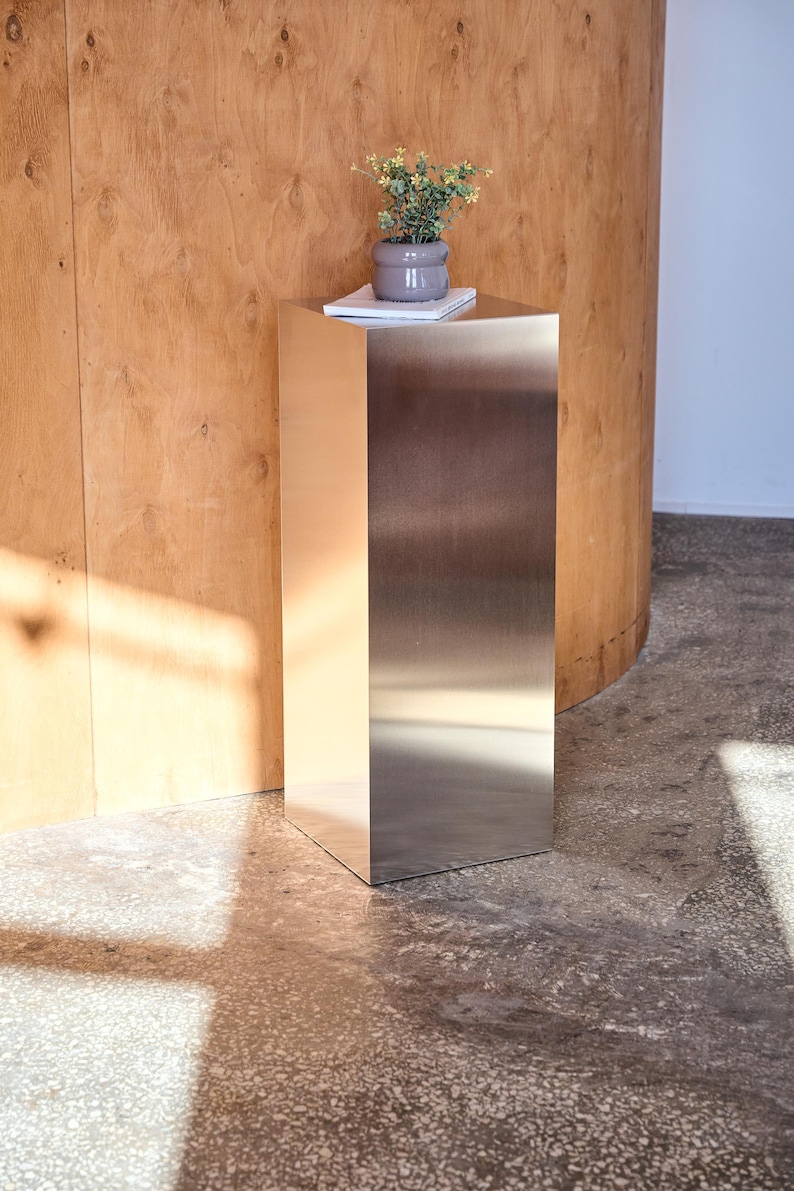 May include: A modern, stainless steel side table with a square top and base. A small, gray ceramic pot with a plant sits on top of the table, resting on a white notepad. The table is in a room with a wooden wall.