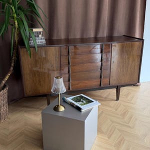 May include: A mid-century modern wooden sideboard with three drawers and two doors. A small, clear glass lamp with a gold base sits on a gray cube table. A magazine is on the table. A tall green plant is in the background.