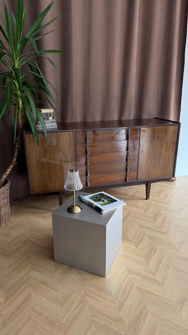 May include: A mid-century modern wooden sideboard with three drawers and two doors. A small, clear glass lamp with a gold base sits on a gray cube table. A magazine is on the table. A tall green plant is in the background.