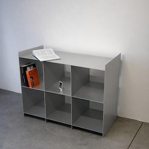 May include: A grey, modular storage unit with six compartments. An open book and a small electronic device sit on top. Two books are visible in one compartment. The unit is made of metal and sits on a concrete floor.