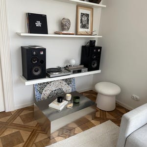 May include: A modern living room setup featuring a stainless steel coffee table, a gray sofa, and a white rug. Shelves display art, vases, and a record player with speakers. A mushroom-shaped stool and decorative candles complete the scene.