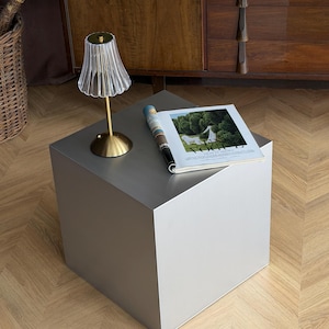 May include: A modern, silver-colored cube-shaped side table with a small lamp and a magazine on top. The lamp has a gold base and a ribbed, clear shade. The magazine is open to a page with a landscape image.