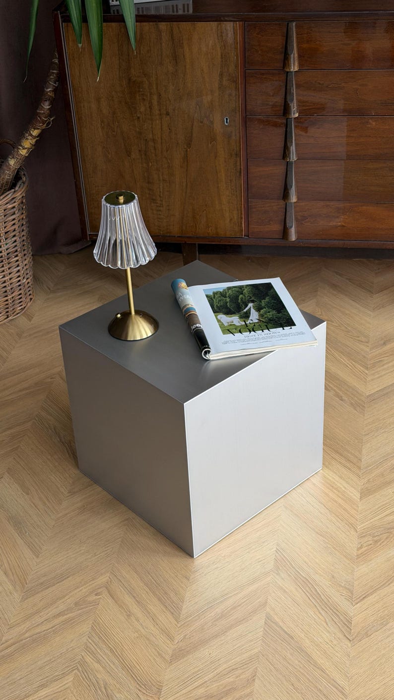 May include: A modern, silver-colored cube-shaped side table with a small lamp and a magazine on top. The lamp has a gold base and a ribbed, clear shade. The magazine is open to a page with a landscape image.