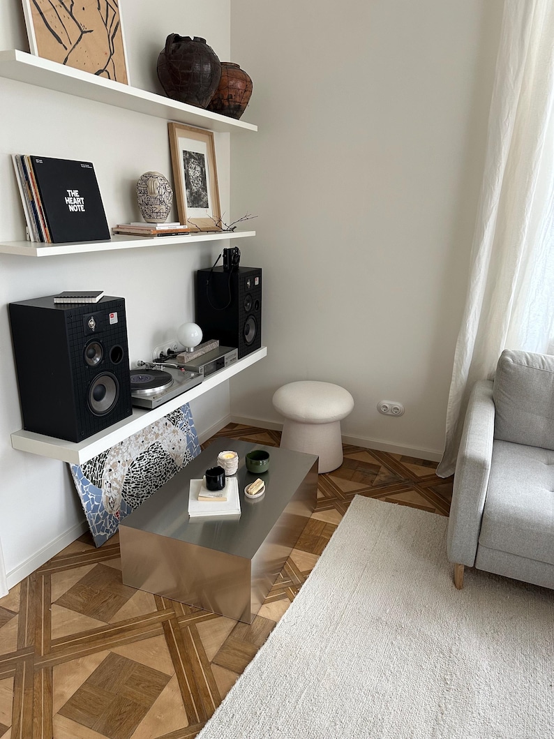 May include: A modern living room with a stainless steel coffee table, a white rug, and a gray sofa. Shelves display art, vinyl records, and speakers. The room has a minimalist aesthetic with a neutral color palette.