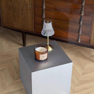 May include: A modern, gray cube-shaped side table with a candle and small lamp. The candle is in a glass jar with a label. The lamp has a clear, fluted shade and a gold base. The table is on a wooden floor.