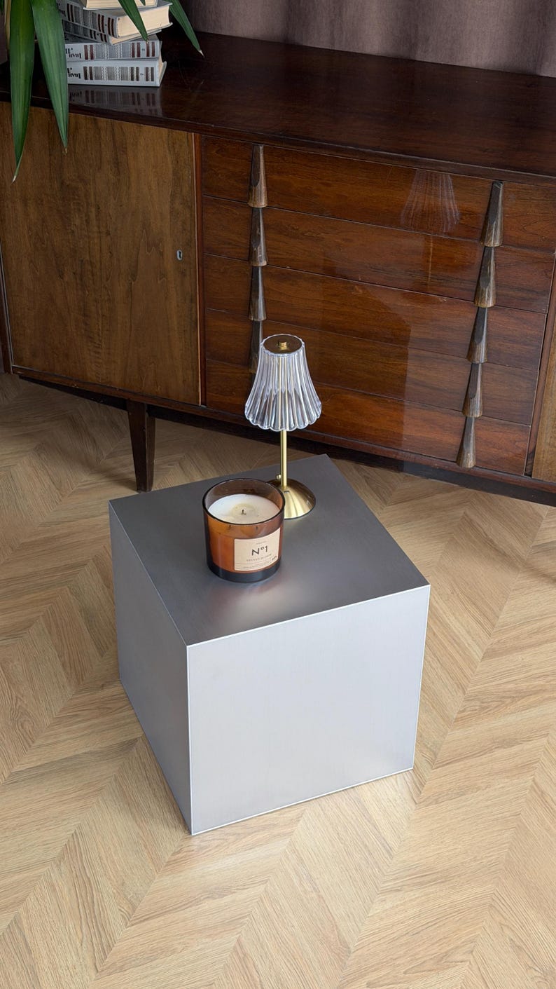 May include: A modern, gray cube-shaped side table with a candle and small lamp. The candle is in a glass jar with a label. The lamp has a clear, fluted shade and a gold base. The table is on a wooden floor.