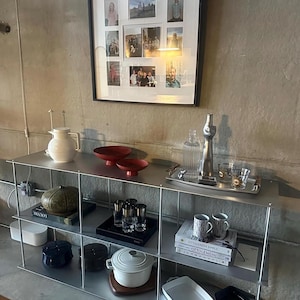 May include: A modern metal shelving unit displays various home decor items. A framed collage of photos hangs above. The shelves hold a white pitcher, red bowls, a decorative bottle, glasses, mugs, books, and other kitchenware.