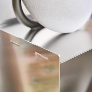 May include: A modern, silver speaker with a rounded, light grey fabric top and a dark grey handle. The speaker sits on a matching silver metal shelf with a minimalist design, showcasing a sleek, contemporary aesthetic.