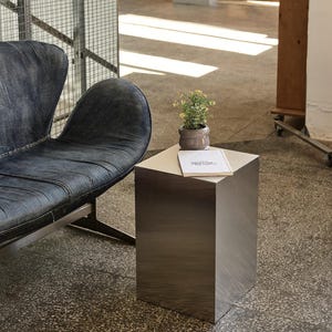 Puede incluir: Escena interior moderna con una mesa cúbica de metal cepillado, una pequeña planta en maceta y un libro. Un sofá de cuero azul oscuro está a la izquierda. El suelo es de hormigón.