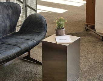 May include: A modern interior scene featuring a brushed metal cube table with a small potted plant and a book on top. A dark blue leather sofa is to the left. The floor is concrete.
