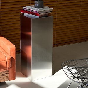 May include: A tall, rectangular metal side table with a brushed finish. The table has a stack of books and a small potted plant on top. The table is next to a leather armchair and a wire chair.