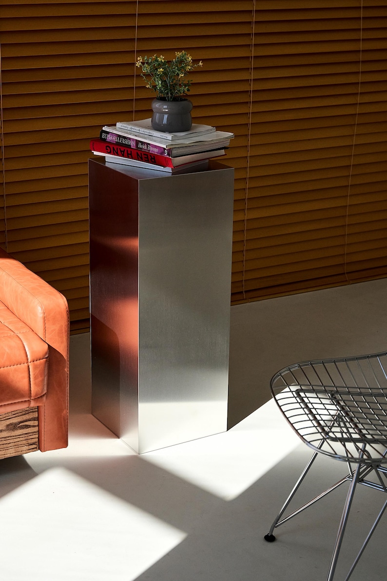 May include: A tall, rectangular metal side table with a brushed finish. The table has a stack of books and a small potted plant on top. The table is next to a leather armchair and a wire chair.