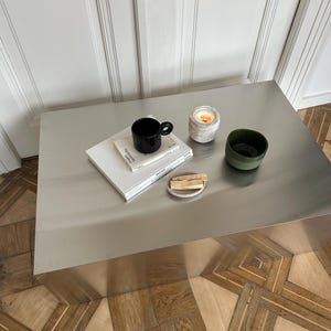 Könnte beinhalten: Ein minimalistischer Couchtisch mit einer schwarzen Tasse, einer kleinen Kerze, einer grünen Schale, Büchern und Holzstäbchen auf einem kleinen Teller. Der Tisch ist aus gebürstetem Metall und steht auf einem Holzboden.