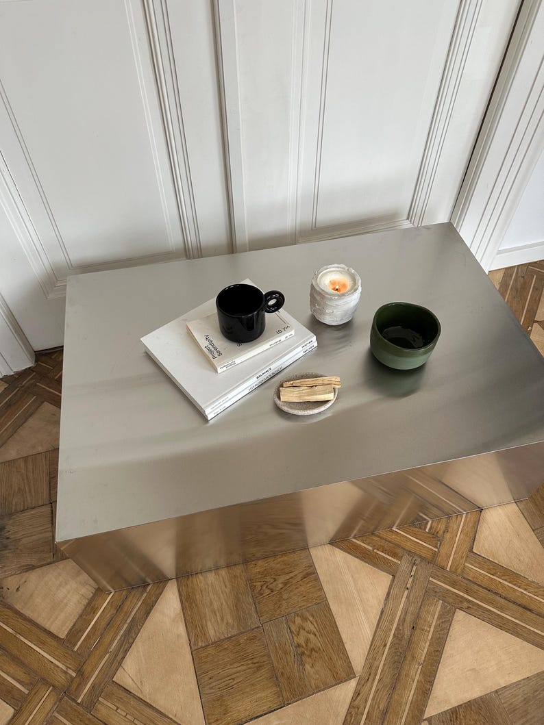 May include: A minimalist coffee table with a black mug, a small candle, a green bowl, a stack of books, and a small plate with wooden sticks. The table is made of brushed metal and sits on a wooden floor.
