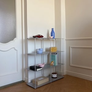 May include: A contemporary metal shelving unit with four tiers, showcasing decorative objects. The top shelf has a red toy car, a blue bottle, and a small pink bowl. Other shelves display books, bowls, a box, and a small decorative item.