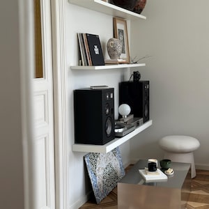 May include: A modern living room corner with white floating shelves displaying decor. Items include black speakers, a record player, framed art, and decorative vases. A silver coffee table and a small stool complete the scene. The floor has a geometric pattern.
