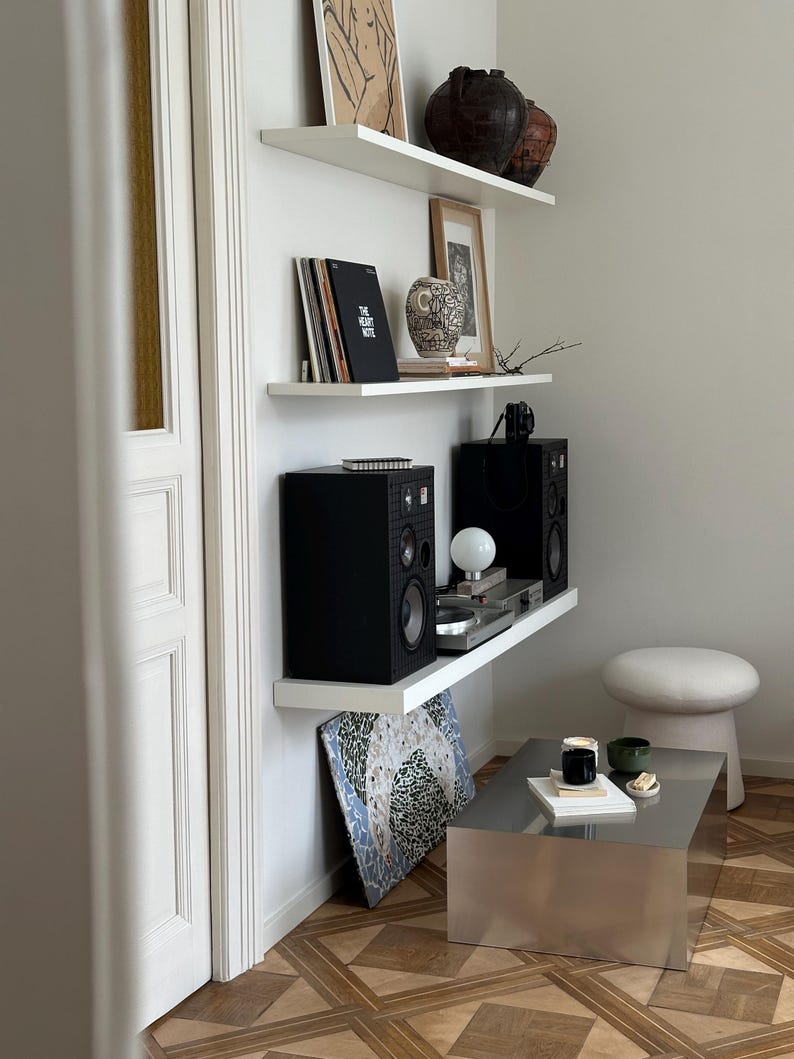 May include: A modern living room corner with white floating shelves displaying decor. Items include black speakers, a record player, framed art, and decorative vases. A silver coffee table and a small stool complete the scene. The floor has a geometric pattern.