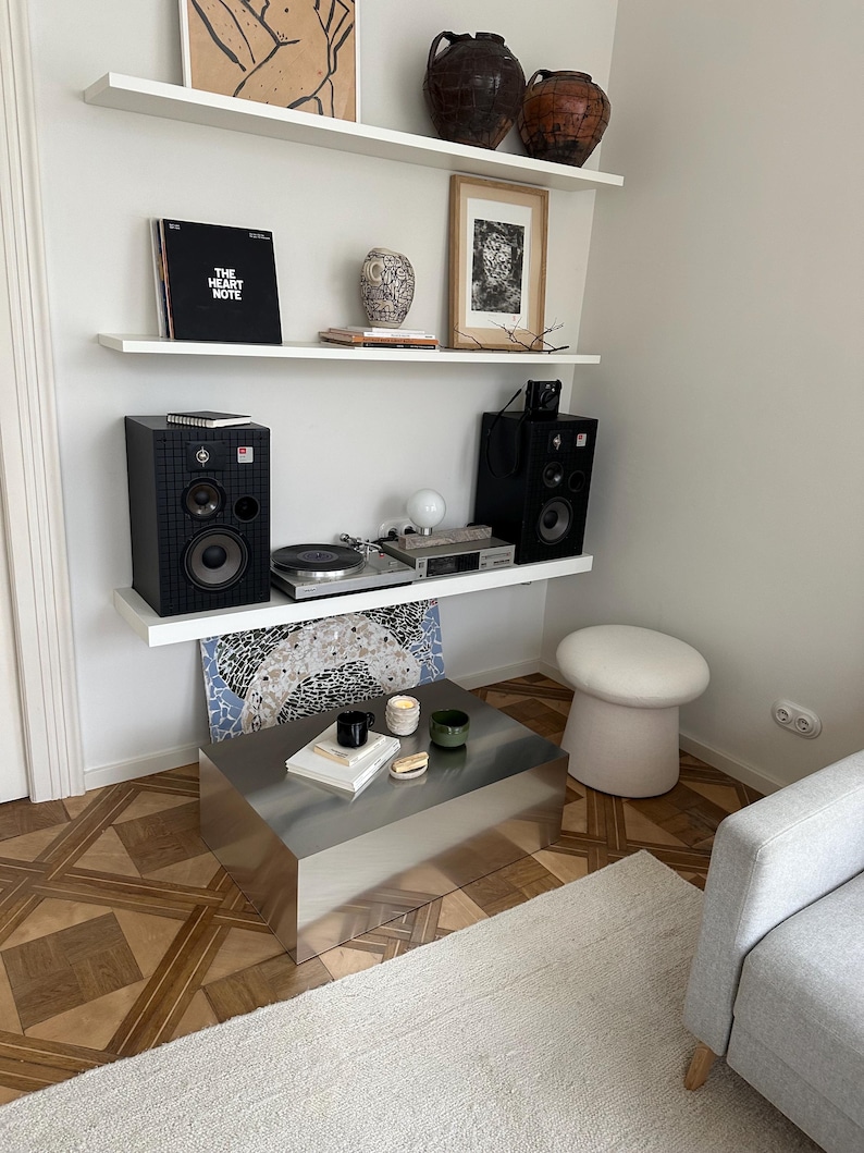 May include: A modern living room setup featuring a stainless steel coffee table, a gray sofa, and a white rug. Shelves display art, vases, and a record player with speakers. A mushroom-shaped stool adds a unique touch.