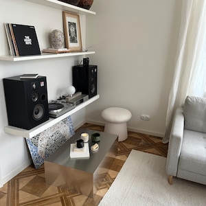 May include: A modern living room with white shelves displaying art, records, and speakers. A silver metal coffee table sits on a patterned wooden floor, with a white rug and a gray sofa. A white stool is next to the table.
