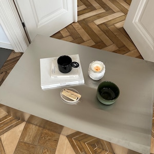 May include: A modern coffee table with various decorative items. A black mug sits atop a stack of books, next to a lit candle in a white holder, a small green bowl, and a small plate with wooden sticks.