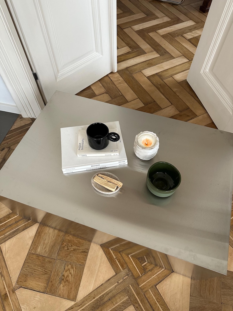 May include: A modern coffee table with various decorative items. A black mug sits atop a stack of books, next to a lit candle in a white holder, a small green bowl, and a small plate with wooden sticks.