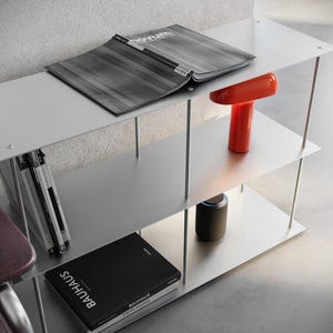 May include: A modern, silver metal shelving unit with three shelves. A magazine, a red lamp, and books are displayed on the shelves. The books include the title "Bauhaus". The unit is supported by thin, silver metal rods.