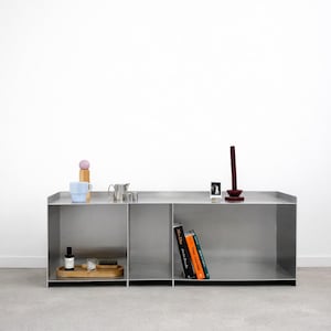 Puede incluir: Un mueble de almacenamiento moderno de metal plateado con tres compartimentos. La superficie superior muestra una pequeña bandeja de madera con un cepillo y una botella, una taza azul, una tetera pequeña y libros. Un objeto decorativo alto y rojo oscuro se encuentra a la derecha.