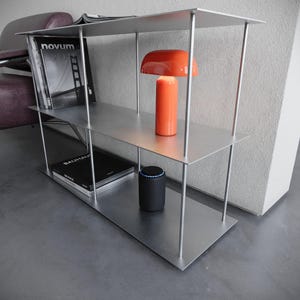 Könnte beinhalten: Modernes Bücherregal aus silberfarbenem Metall mit drei Regalböden. Auf dem mittleren Regal steht eine orangefarbene Tischlampe, auf den anderen Regalböden Bücher und ein intelligenter Lautsprecher. Das Regal wird von dünnen, vertikalen Metallstangen getragen.
