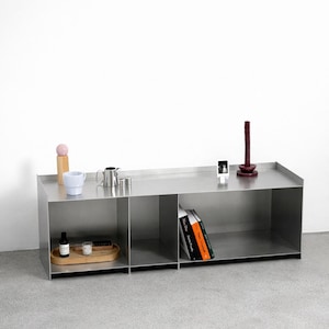May include: A modern, stainless steel storage unit with open shelving. The unit holds books, decorative items, and a wooden tray with small objects. A tall, dark red candle sits on top.