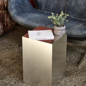 May include: A contemporary side table with a brushed metal finish. A small potted plant and a book rest on the table's surface. The table is placed on a speckled floor, with a denim chair in the background.