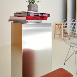 May include: A modern interior scene featuring a metallic pedestal table topped with books and a small potted plant. The books have red and white covers. A wire chair and a leather sofa are also visible.