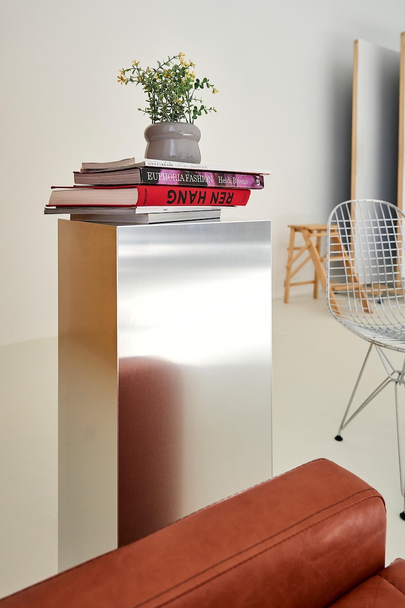 May include: A modern interior scene featuring a metallic pedestal table topped with books and a small potted plant. The books have red and white covers. A wire chair and a leather sofa are also visible.