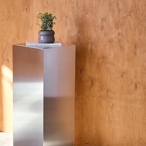 May include: A brushed metal pedestal with a small potted plant and a book on top. The plant has green leaves and is in a gray pot. The pedestal is rectangular and stands against a wood-paneled wall.