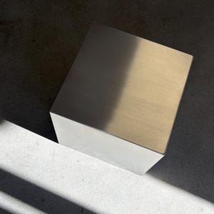 Puede incluir: Un objeto cúbico sólido con un acabado metálico cepillado suave. La superficie superior refleja la luz, creando un contraste marcado entre un lado brillante e iluminado y un lado más oscuro y sombreado, resaltando su forma geométrica.