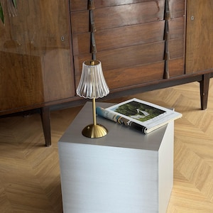 May include: A modern side table with a brushed metal finish. A small lamp with a gold base and a fluted glass shade sits on top, next to a magazine. A dark wood cabinet is in the background.