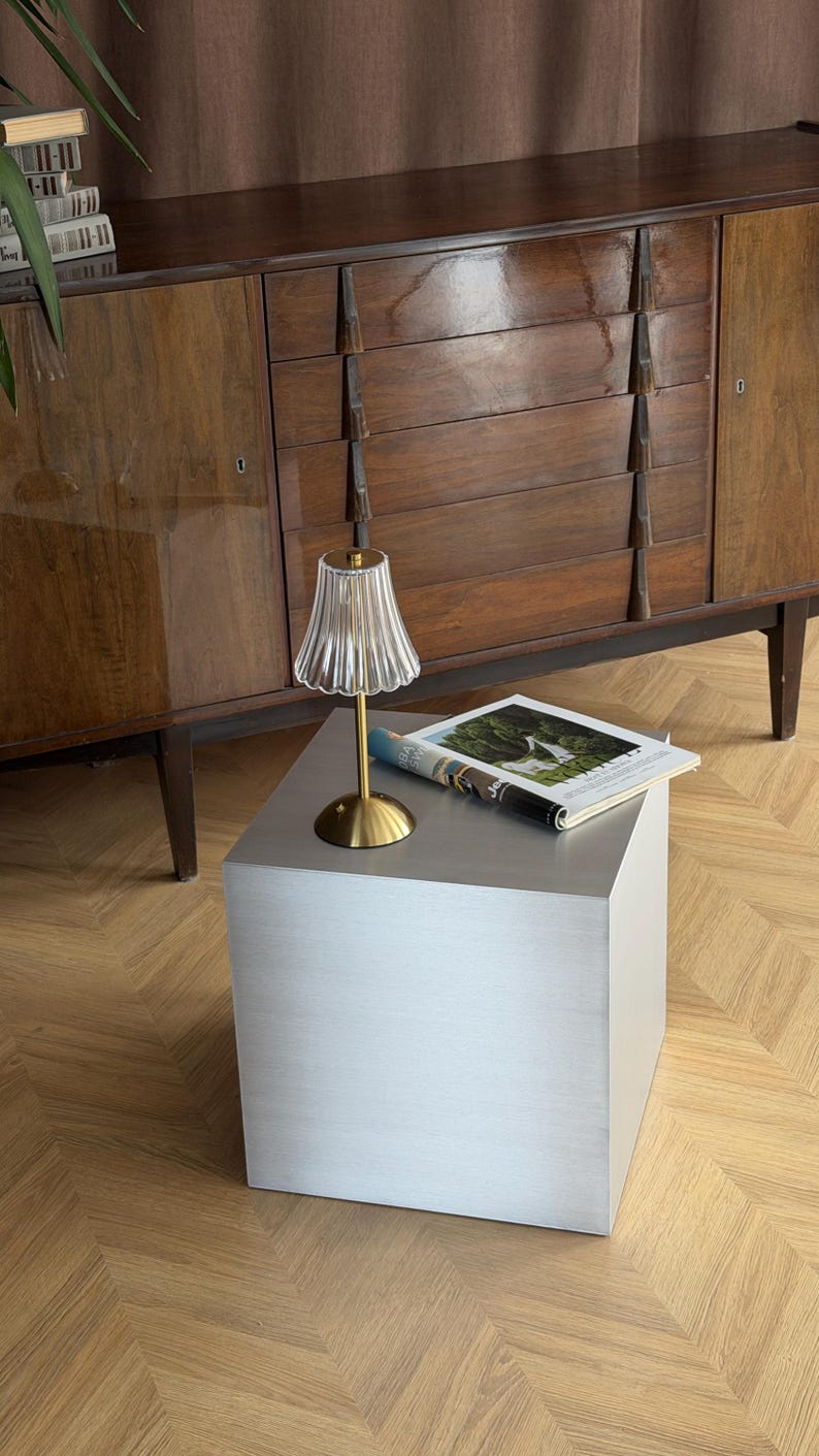May include: A modern side table with a brushed metal finish. A small lamp with a gold base and a fluted glass shade sits on top, next to a magazine. A dark wood cabinet is in the background.